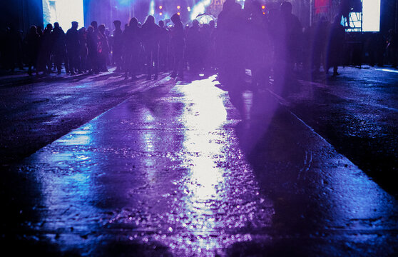 A Crowd Of People Stands On Wet Pavement In Front Of The Stage At A Concert. Light From Event Is Beautifully Reflected