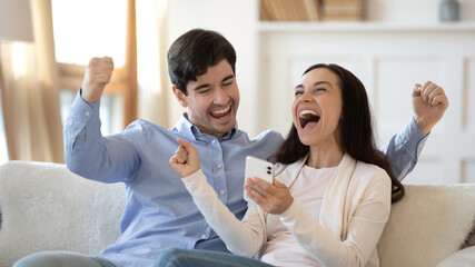 Excited young couple triumph winning online lottery on smartphone gadget, overjoyed man and woman relax at home feel euphoric get read unexpected great news or sale offer on internet on cellphone