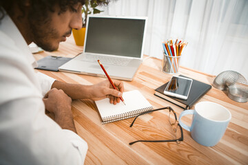 Young serious millennial takes notes with pencil in work notebook while sitting at wooden table...