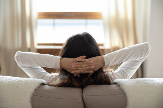 Close up back view of calm woman sit relax on comfortable sofa hands over head, lazy female rest on couch in living room, look in window distance thinking or dreaming, stress free, relaxation concept