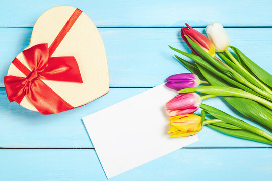 Colorful spring tulip flowers with decorative giftbox and blank photo on light blue wooden background as greeting card. Mothersday or spring concept.