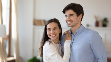 Smiling young caucasian couple renters stand hugging cuddling look in distance dreaming or visualizing, happy man and woman embrace imagine plan bright future together, new beginning concept