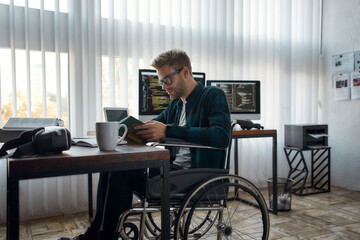Obraz na płótnie Canvas Focused male web developer in a wheelchair reading a book while sitting at his workplace with multiple computer screens on the background