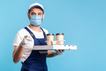 Fototapeta na wymiar Delivery service. Smiling happy courier in overalls and mask holding coffee, pizza box, wearing safety gloves and offering drinks food, pointing finger to camera, choosing you. indoor shot, isolated