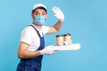Fototapeta na wymiar Delivery service. Amiable courier in overalls and mask holding coffee and pizza box, wearing safety gloves offering drinks food and waving hello to camera. indoor shot isolated on blue background