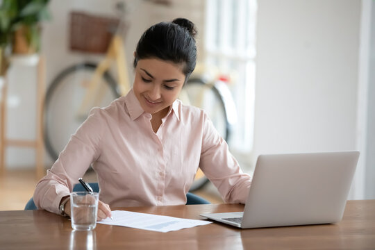 Smiling young indian woman sitting at table with computer, checking economic paper report, making remarks at workplace. Happy hindu businesswoman financial advisor working on project alone in office.