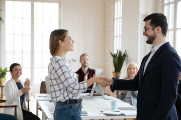 Side view happy young female worker shaking hands with smiling confident boss in eyewear. Satisfied with good job results team leader praising employee at meeting while diverse coworkers applauding.