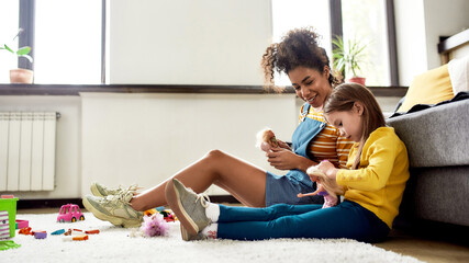 Kidspace. African american woman baby sitter entertaining caucasian cute little girl. They are playing with dolls, sitting on the floor - obrazy, fototapety, plakaty