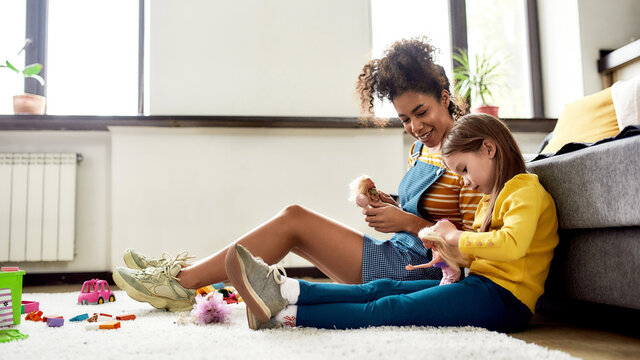 Kidspace. African american woman baby sitter entertaining caucasian cute little girl. They are playing with dolls, sitting on the floor