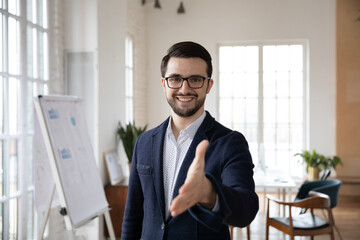 Head shot pleasant smiling trustful manager in suit reaching out hand, looking at camera. Happy confident young businessman welcoming greeting clients partners in modern office, acquaintance concept.