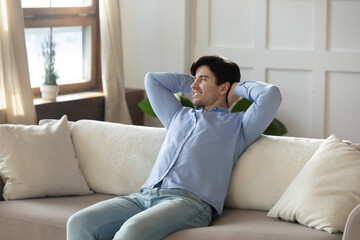 Smiling young caucasian man sit relax on sofa in living room look in window distance dreaming or thinking, happy millennial male rest on couch at home, enjoy leisure weekend, stress free concept