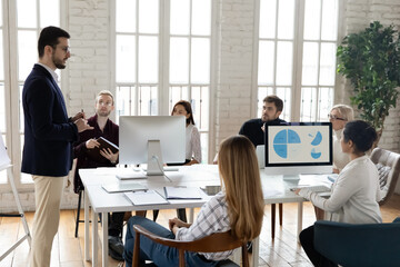 Side view confident male speaker team leader in glasses explaining marketing strategy to multiracial colleagues. Focused young diverse teammates sitting at shared table, attending workshop in office.