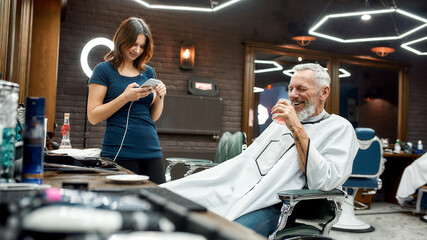 Barbershop is not just a haircut. Handsome middle-aged man drinking something and having fun while sitting in armchair and talking with young girl barber girl