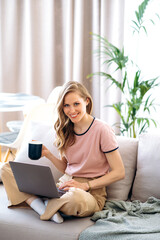 Beautiful caucasian blue-eyed woman with a charming smile dressed in stylish casual clothes drinks a coffee while sitting on a sofa with a laptop at home