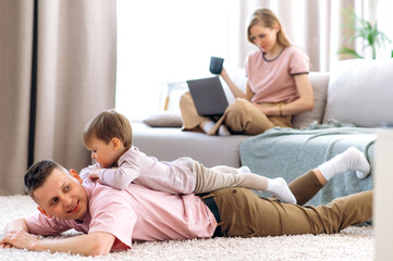 Fototapeta premium Young dad spends time with his little son, they lie together on the floor and smile, and mom in the background sits on the couch with a laptop and shopping online