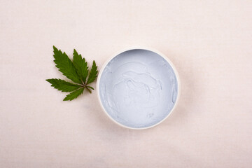 white round jar with a body scrub with a leaf of marijuana on a white background top view, cosmetics, skin care, beauty