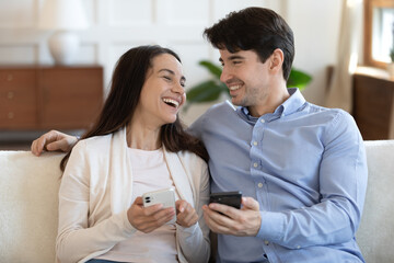 Overjoyed young caucasian couple relax on sofa on weekend at home have fun using smartphones...