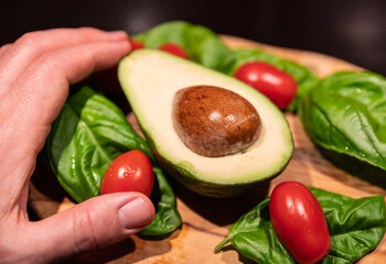 A wooden cutting board on a black background with an avocado in the center. All around, intense green basil leaves and bright red cherry tomatoes.  One hand touches the avocado.
