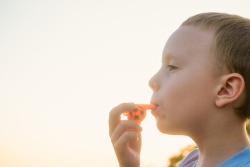 Little boy blowing in orange color whistle on a bright background of evening sunny sky