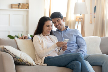 Overjoyed young caucasian man and woman sit rest on couch in living room have fun using smartphone together, happy millennial couple laugh watching funny video on cellphone, enjoy weekend indoors