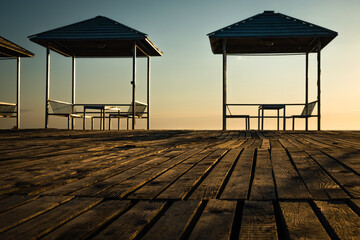 Empty summer cafe on the beach. No visitors due to pandemic and covid-19. Tables on a canopy, wooden flooring. Restaurant with sea view on the coastline. Closed borders, lack of tourists. Evening. Sun