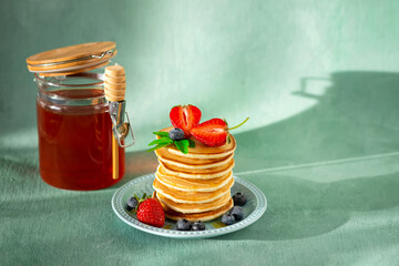 Stack of pancakes with strawberries, blueberries and jar of honey on green background.