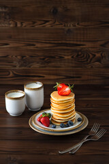 Stack of homemade classical pancakes with fresh blueberries and strawberries on plate, fork and two cups of milk on wooden background. Vertical orientation, copy space. Delicious sweet dessert.