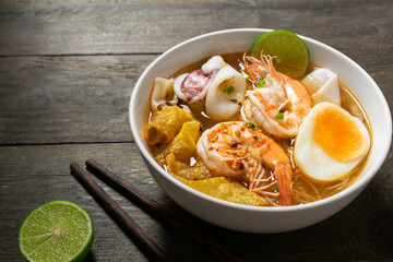 Spicy rice noodle with seafood and crispy wonton in white bowl on wooden table.