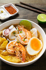 Spicy rice noodle with seafood and crispy wonton in white bowl on wooden table.