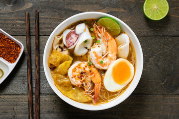 Spicy rice noodle with seafood and crispy wonton in white bowl on wooden table.