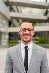 Face of happy Hispanic bald bearded businessman with eyeglasses smiling in the city outdoors