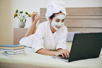 Cheerful young woman with face mask lying on sofa and doing homework on laptop after reading books