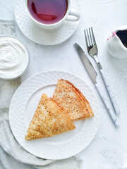 Two thin pancakes on a white plate, tea, sour cream and jam served for Breakfast on a light table top view.