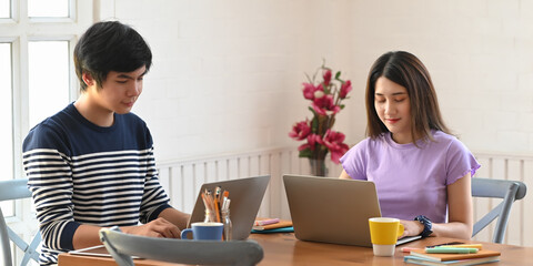 Young couple working with computer laptop and tablet while sitting together at the wooden working desk over comfortable sitting room as background.