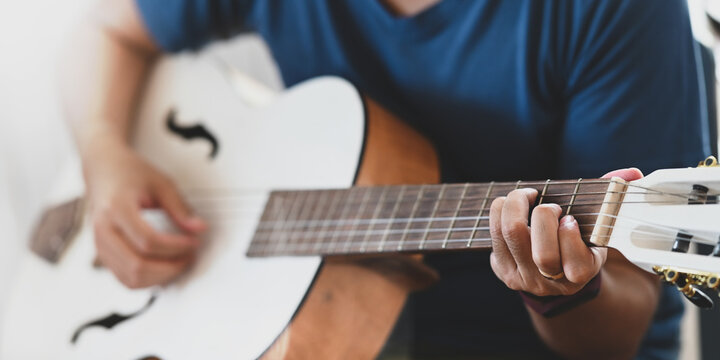 Cropped image of young man practicing on acoustic guitar while sitting over sitting room as background. Man with performing an acoustic guitar concept.
