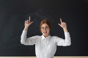 Beautiful smart teacher in glasses is standing in front of the blackboard. There are glasses on her face. She raised her hands up. The index fingers of the girl show yes. The blackboard is clean.