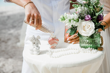 Bride and groom pouring colorful different colored sands into the crystal vase close up during symbolic nautical decor destination wedding marriage ceremony on sandy beach in front of the ocean 