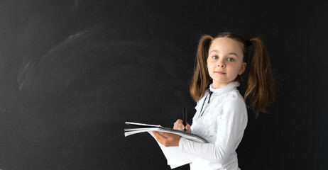 Young student in the office. She stands by a clean and black board and holds a notebook in her hands. Written answers. The girl is looking at the camera and smiling. She is wearing a school uniform.