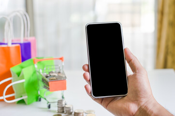 Hand holding mobile phone and In a small cart with paper bag and coin have on it online shopping located in home concept of detention at home to prevent the Covid19 virus.