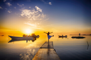 Young woman in white dress dancing on concrete pier over colorful morning sunrise clouds sky with boats floating on water with reflection background, Bahrain.