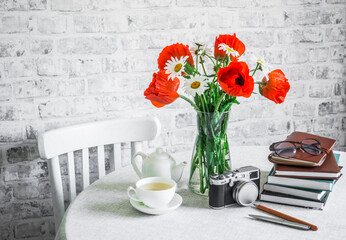 Bunch of poppies, stack of books, cup of green tea, vintage camera on a table in a bright room. Cozy home interior concept