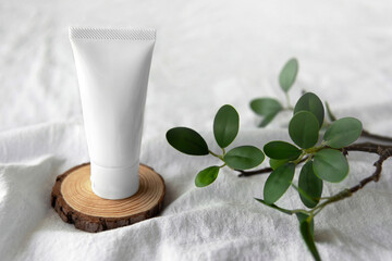 Fototapeta na wymiar Empty organic facial skincare product white tube on wood platform next to branch with leaves on soft white fabric cloth background