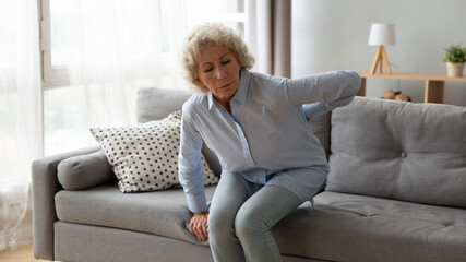 Old woman gets up off the sofa feels shooting repeated low back pain caused by spinal injury degeneration, ruptured discs, skeletal irregularities, arthritis or osteoarthritis chronic diseases concept - obrazy, fototapety, plakaty