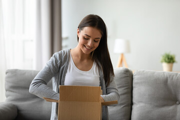 Satisfied client young beautiful woman seated on couch in living room holding cardboard on lap feels pleased of delivery service unpacking box smiling enjoy moment of unbox purchased goods in web shop