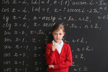 Cute little schoolgirl with raised index finger near blackboard in classroom