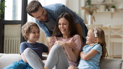 Overjoyed young family with small children have fun playing together at home on leisure weekend, happy father and little daughter and son tickle excited mom, feeling playful engaged in funny game