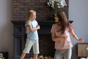 Happy young mother dance together with excited little preschooler daughter, enjoy family weekend at home, smiling millennial mom or nanny have fun play with overjoyed small girl child in living room