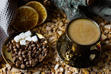 Aromatic dark coffee, with milk with bubbles on the surface in a dark gold Cup on a saucer, next to whole grains, peeled and dried oranges. Background of alder chips and wood snags in the background