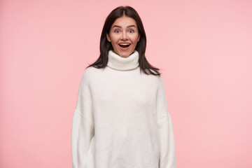 Surprised young lovely brunette female with loose hair looking excitedly at camera with opened mouth and keeping hands along body while posing over pink background