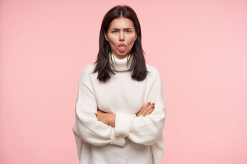 Fototapeta premium Offended young brown haired woman with casual hairstyle keeping her hands crossed and showing tongue to camera while standing against pink background